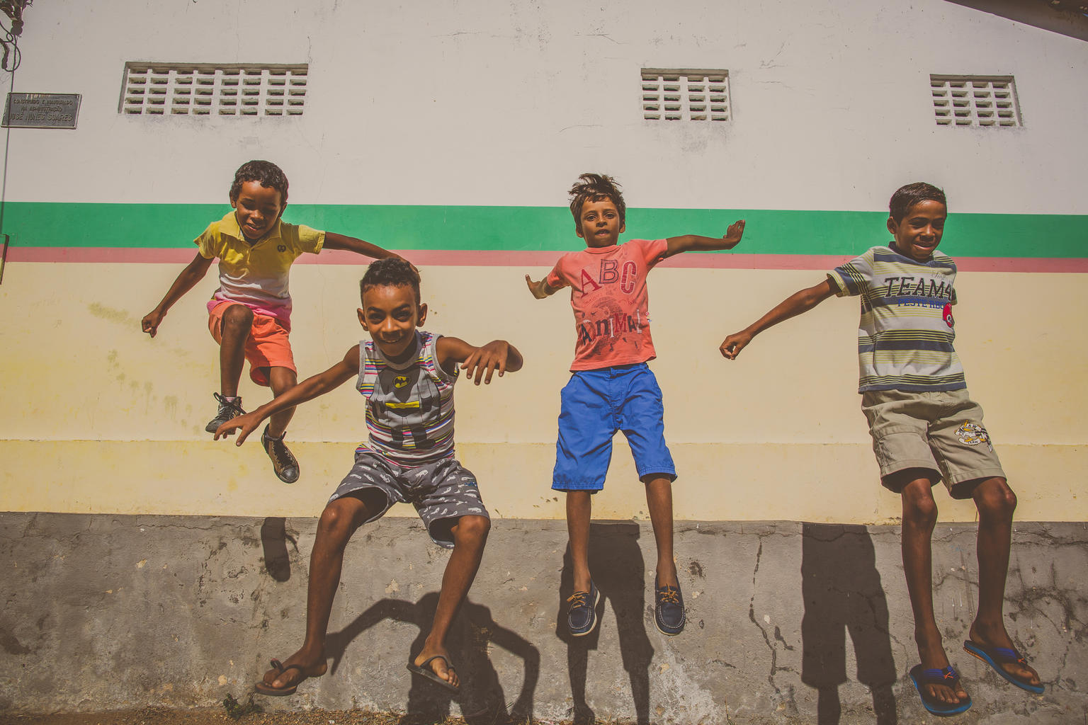 Quatro meninos pulam enquanto olham para a câmera. No fundo, a parede de uma escola
