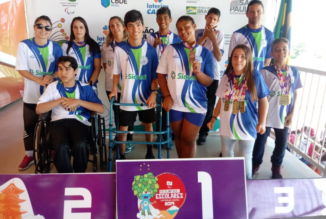 A imagem mostra 11 pessoas, adolescentes e adultos, em um pódio. Todos usam o mesmo uniforme e alguns adolescentes seguram medalhas em suas mãos.