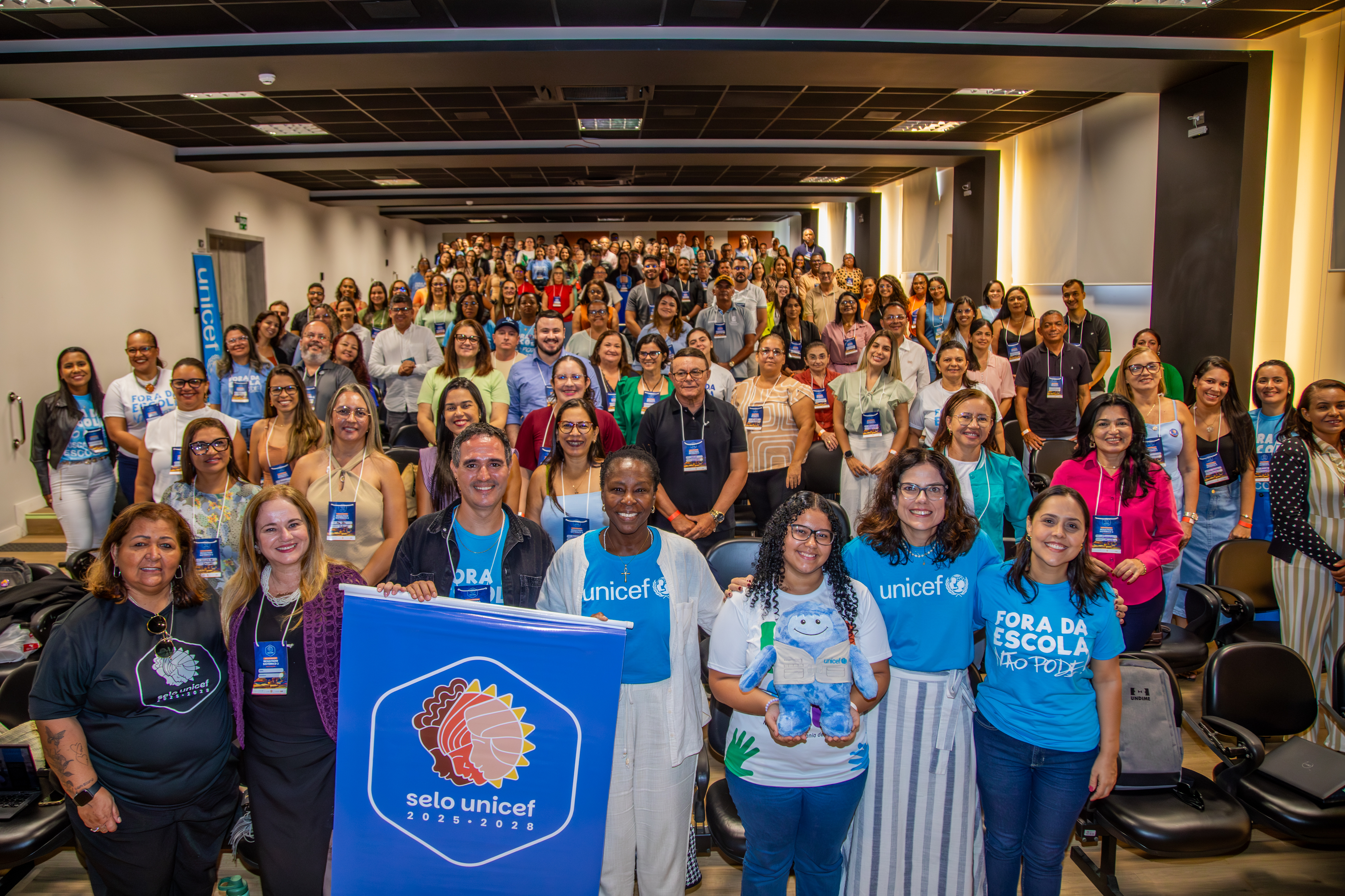 Grupo de participantes da formação, cerca de 200 pessoas, pousam para foto dentro do auditório com o mascote Unicefito