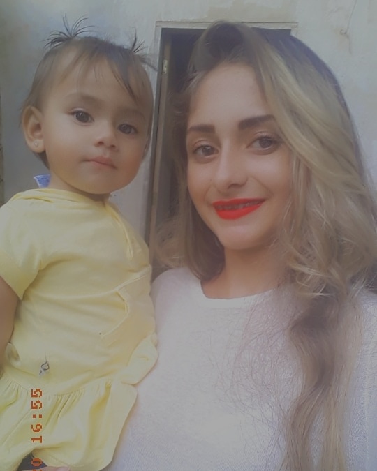Jovem de cabelo loiro e vestindo uma blusa branca segura a filha no colo. A bebê menor de 1 ano veste um vestidinho amarelo e olha para a frente
