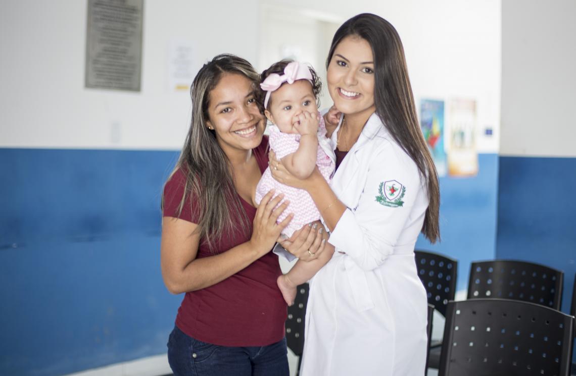 A imagem mostra Maria de Fátima (mãe) e uma médica, que utiliza jaleco branco, segurando Júlia (filha), que está entre as duas mulheres. As três estão em uma recepção de um ambiente hospitalar.