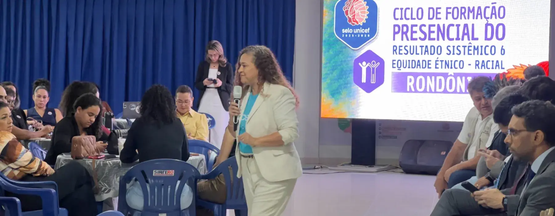 mulher usa blusa do UNICEF enquanto palestra e caminha no meio de pessoas sentadas