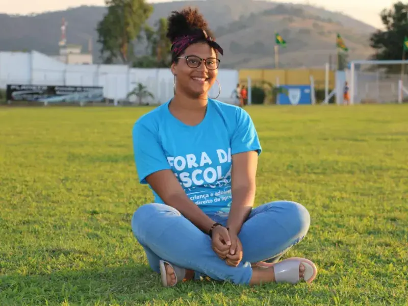 Adolescente sentada num gramado verde sorri para câmera. Ela veste calça jeans azul e uma blusa azul. Óculos de grau redondos, brincos de argola e uma faixa roxa e preta no cabelo 