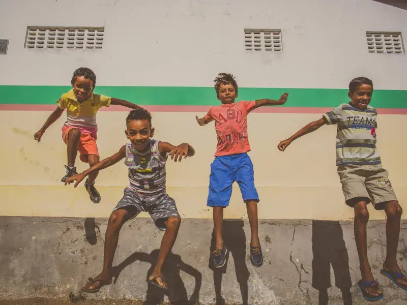 Quatro meninos pulam enquanto olham para a câmera. No fundo, a parede de uma escola