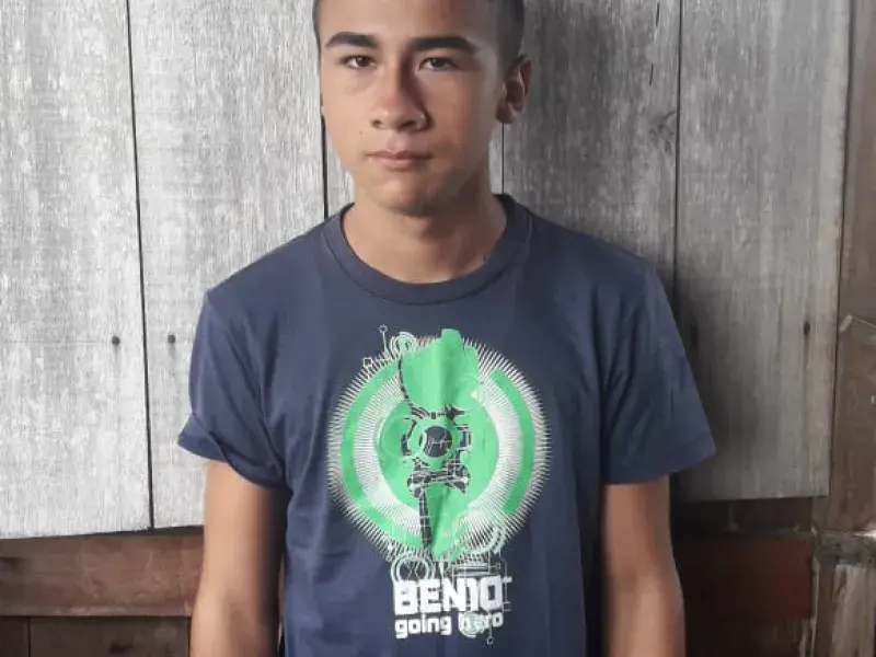 Adolescente em pé, veste camiseta azul marinho com estampa verde fosforescente. Nos pulsos, um relógio verde e pulseiras artesanais