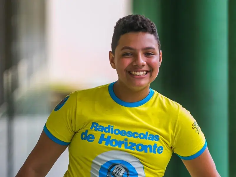 Adolescente sorrindo olha para a câmera. Veste camiseta amarela fosforescente com detalhes em azul na manga. A estampa diz: "Radioescolas de Horizonte"  