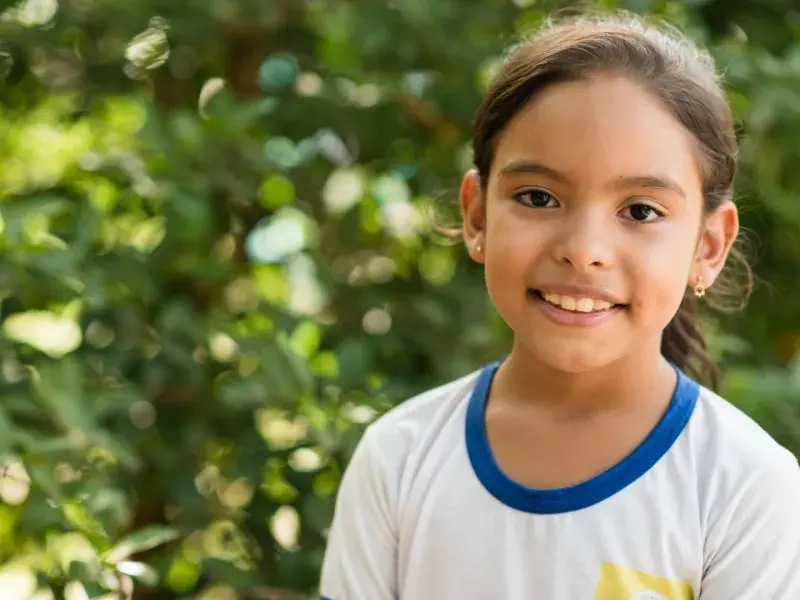 Criança com uniforme escolar branco de gola azul olha para a câmera e sorri. Ao fundo, temos uma vegetação verde