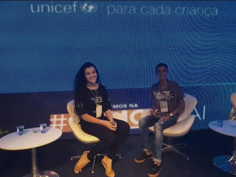 Adolescente vestindo blusa e calça preta sorri. Ela segura um microfone nas mãos e está sentada numa cadeira em cima de um palco. ao fundo, um painel do UNICEF. Ao lado dela, outro jovem sentado numa cadeira sorri