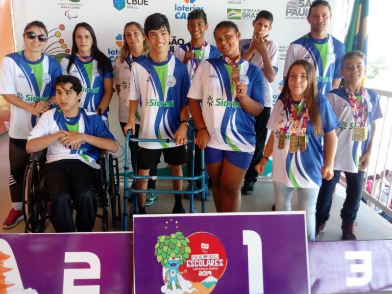 A imagem mostra 11 pessoas, adolescentes e adultos, em um pódio. Todos usam o mesmo uniforme e alguns adolescentes seguram medalhas em suas mãos.