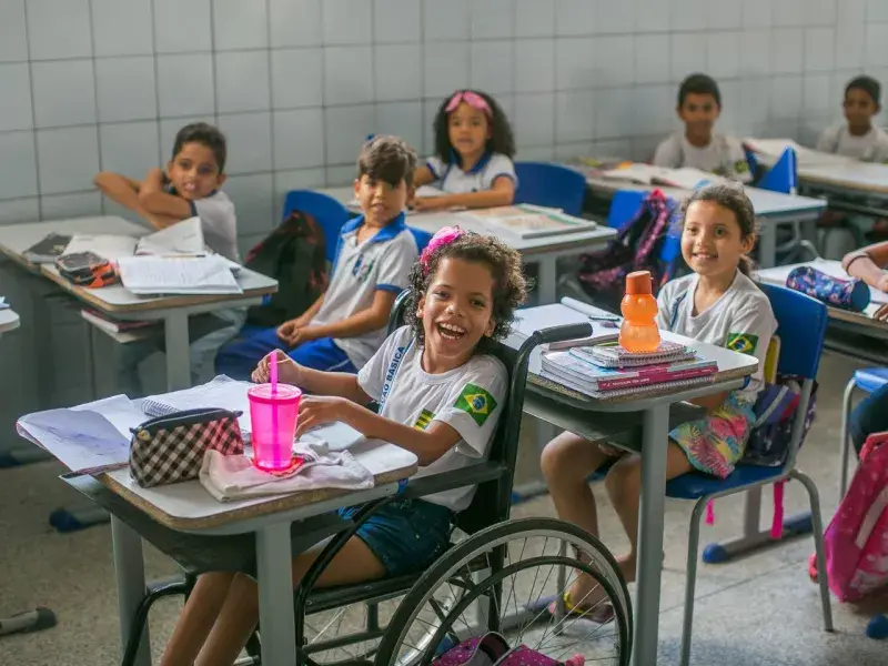 Vestindo uniforme escolar, menina sorri para câmera. Ela está numa cadeira de rodas encaixada na carteira escolar. Sobre a mesa, um estojo quadriculado preto e branco, uma garrafinha de água rosa e um caderno. Ao fundo da menina, outras crianças estão sentadas em suas carteiras vestindo uniforme escolar e olhando para frente