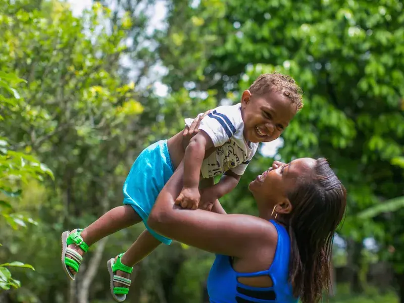 Mulher quilombola veste blusa azul e sorri para o filho pequeno. Ela o ergue. O menininho sorri, suspenso no ar. Ele veste uma blusa branca com listras azuis escuras e um desenho amarelo no peito, bermuda azul claro e sandalhas verdes fosforescentes. Ao fundo, uma vegetação verde