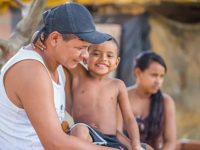 Na foto, um menino indígena está sentado no colo de seu pai e sorrindo. Seu pai, de blusa branca e boné, também sorri. Ao fundo, de forma desfocada, é possível ver uma mulher e residências.