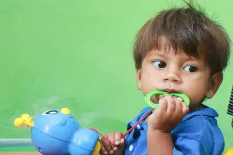 Bebê de um ano vestindo camiseta azul, olha para o lado enquanto mastiga o aro verde de um brinquedo azul