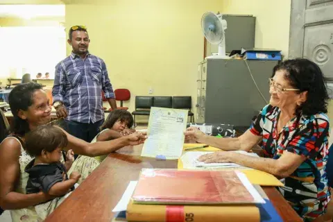 Mulher sentada com o filho no colo olha para a conselheira tutelar sentada a sua frente. A mulher recebe o registro civil dos filhos. A cabeça da filha repousa no tampo da mesa, As duas mulheres seguram o documento sobre a mesa de madeira. Ao fundo, um funcionário municipal assiste à cena 