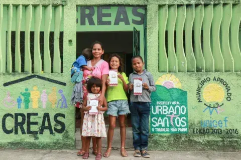 Mulher com vestido rosa segura um bebê no colo. O bebê está virado de costas para a câmera. Na frente e ao lado da mãe, seus outros 3 filhos olham para a câmera. As crianças seguram nas mãos um panfleto do governo municipal. Eles estão na porta de um CREAS, o Centro de Referência em Assistência Social. No muro verde do edifício, estão pintados o logo do CREAS, do município de Urbano Santos e do Selo UNICEF