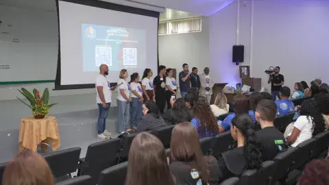 Foto de trás do auditório com várias pessoas sentadas e outras em pé apresentando