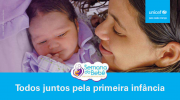 Uma mãe, sorrindo, olha para sua filha bebê. Na imagem está escrito Todos Juntos Pela Primeira Infância.