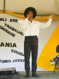 Jovem vestindo camisa branca e calça preta fala ao microfone em cima de um palco. Ele está com os braços abertos e segura em uma das mãos um livro